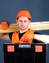 Man in helmet, hard hat holds toolbox and wooden beams, grey background. Professional woodworker concept. Carpenter