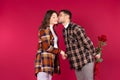 A man hiding a bouquet of red roses behind his back kissing his girlfriend on the cheek when meeting. Red background