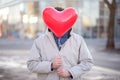 Man hiding face behind red heart-shaped air balloon. Guy standing in city street with covered face. Blind date concept Royalty Free Stock Photo