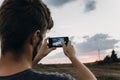 man hipster traveler taking photo holding smart phone, of beautiful sunset scenery in summer field. instagram photography.