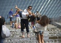 Man holding laughing girl on hands standing among water jets of the fountain, another girl taking picture