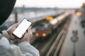 The Man is holding a mock up smartphone in his hand, against the background of the train at the railway station. Royalty Free Stock Photo