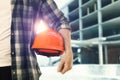 Man holding orange hard hat at construction site with unfinished building, closeup. Space for text Royalty Free Stock Photo