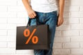 Man holding paper shopping bag with percent sign near white brick wall, closeup. Discount concept