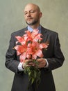 Man holding pink lily bouquet in his hands. Model is bald with grey beard, wearing classic grey suit. Handsome male with special
