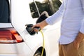 Man holding power supply cable at electric vehicle charging station. Hansome guy holding plug of the charger, while car