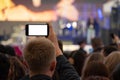A man is holding a smartphone in his hands and is recording or live broadcasting a street concert among a crowd of fans. View from