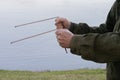 man holding two steel stick together near water with grass and trees in background