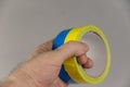 A man is holding yellow and blue duct tape in his hand. Reel of colored duct tape against the gray background. Selective focus. No