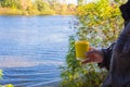 Man holding yellow cup on river background. Coffee cup in nand on autumn landscape background. Travel concept. Vacations at nature