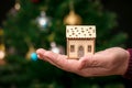 A man holds in his hand a wooden toy house near the Christmas tree. House as a gift for the New Year