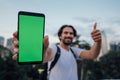 A man holds a phone with a green screen and stands against the backdrop of a metropolis