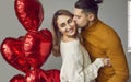Man hugs his woman, kisses her on the cheek and gives her red heart shaped Valentine balloons Royalty Free Stock Photo