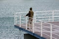 Man in hunting jacket and hoodie standing at the end of local pier fishing with small fishing rod over white metal fence Royalty Free Stock Photo