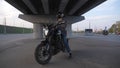 A man in a jacket and helmet sits on an electric motorcycle under a bridge. Retracting camera.