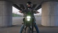 A man in a jacket and helmet sits on an electric motorcycle under a bridge. Retracting camera.