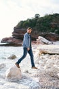 Man in jeans and a denim jacket takes a step on a pebble beach from a large stone against a background of green rocky