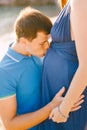 Man kissing pregnant belly of woman in long blue dress on sunny day