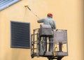 Man on a lifting platform painting the building wall with a roller exterior outdoors. Worker on a ladder manually painting yellow