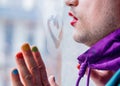 Man with lipstick and polish nail stay near misted window with drawing heart