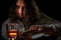 Man with long hair reading an old book in dark room. Portrait of a caucasian white male in medieval clothes and a glass of drink,