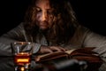 Man with long hair reading an old book in dark room. Portrait of a caucasian white male in medieval clothes and a glass of drink,