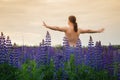 Man with long hair stands with his arms wide spread on green field with purple lupines