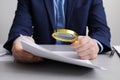 Man looking at document through magnifier at white table, closeup. Searching concept