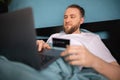 Man lying in bed doing online shopping using credit card and laptop while relaxing in bed on weekend Royalty Free Stock Photo