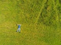 Man lying on the ground in a park on a green grass field. Man dressed in blue t shirt and jeans hands behind hes head. Aerial top