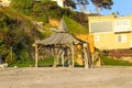 A man made wooden hut on a sandy beach near a yellow house and a lush green hillside Royalty Free Stock Photo