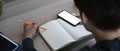 A man making note on empty notebook and using mobile phone on wooden table. Royalty Free Stock Photo