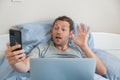 Man making video call on smartlphone waving hello communicating online while lying in bed with laptop in a bed at home