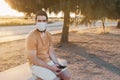 Man in a mask sitting on a bench and texting someone with his smart phone. He is wearing a yellow t-shirt and a white pants Royalty Free Stock Photo