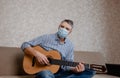 a man in a medical mask plays guitar at home on the couch. Home entertainment in isolation Royalty Free Stock Photo