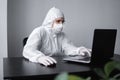 Man in medical mask and protective suit, rubber gloves sits at home and works at laptop at the table during quarantine Royalty Free Stock Photo