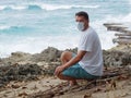 A man with a medical mask setting alone on the reef beach at summer day. Tropical island, coastline.