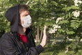 Man in a medical mask wearing a black hood shows Stop gesture to a tree flowers.