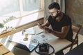 Man millenial in glasses sits at table in front of laptop and uses smartphone, reads message from business partner