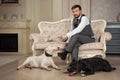 Man with mobile phone seating on the sofa. Dogs: black pit bull or stafforshire terrier, white bull terrier seatting in the legs o