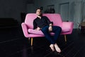 Man model sits on pink sofa in loft interior. Businessman in a black shirt and watch, blue pants.