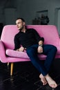 Man model sits on pink sofa in loft interior. Businessman in a black shirt and watch, blue pants. Royalty Free Stock Photo