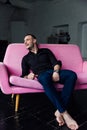 Man model sits on pink sofa in loft interior. Businessman in a black shirt and watch, blue pants.