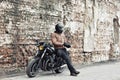Man a motorcyclist standing with helmet near her bike, brick wall of garage background. Male in a leather jacket and