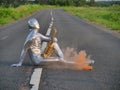 Man musician saxophonist in full body-hugging silver and silver electric suit holding golden alto saxophone, sitting on empty road