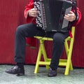 Man in national Russian shirt sits on chair and plays accordion against dark red wall.