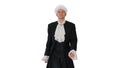 Man in old-fashioned laced frock coat and white wig walking in a mannered way looking at camera on white background.