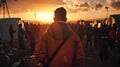 A man in an orange jacket stands in front of a crowd of people in a refugee camp. The sun is setting in the background