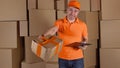 Man in orange uniform delivering heavily damaged parcel to customer. Brown cartons background. Unprofessional work and