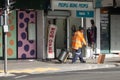 Man in orange working jacket pulling bin on small cart passes under shop sign People Being People